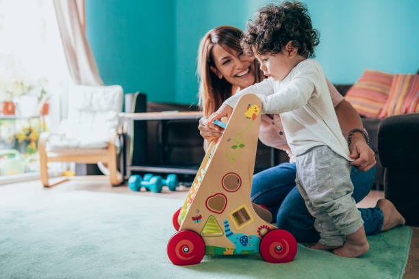 Mother is playing with her cute little baby boy on the floor with walker Un joli petit garçon jouant avec son chariot en bois, accompagné de sa maman